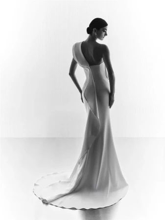 Model in a white one-shoulder gown with a fluted train standing against a seamless white studio background.