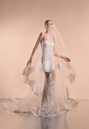 Model Mili Boskovic in a strapless lace wedding dress with a sheer cathedral veil over her head against a warm pleated studio backdrop.
