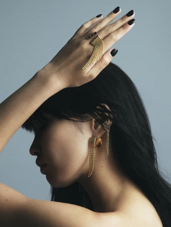Portrait shot of a woman showcasing golden jewelry, including a ring and earrings, against a gray studio backdrop with dark hair.