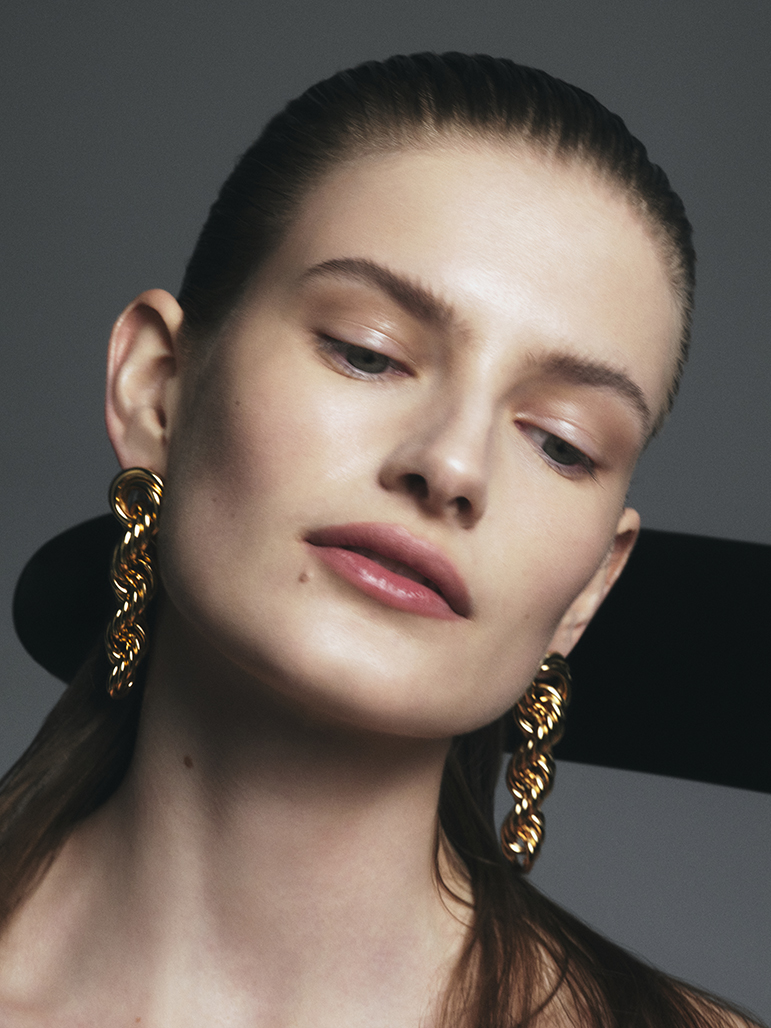 Close-up portrait of a fair-skinned adult woman with gold chain earrings against a grey background and black geometric shape.