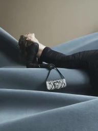 Model in a black dress reclining on a blue sculpted set holding a snakeskin print shoulder bag