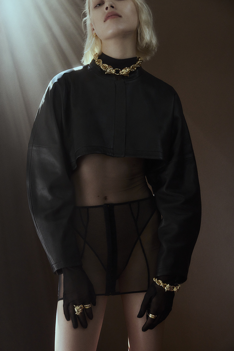 Model wearing a black crop top and sheer black skirt with gold chain necklace and bracelet against a dark background