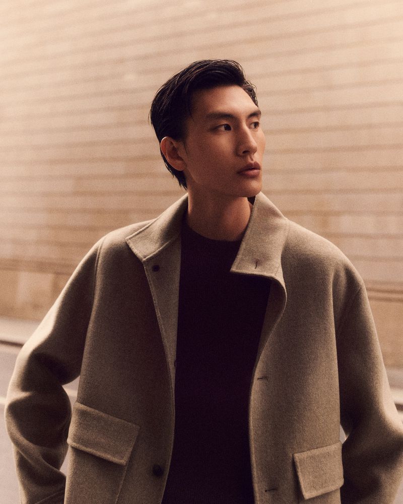 Portrait of a young adult male wearing a beige wool coat and a dark sweater, looking to the side in a studio environment.