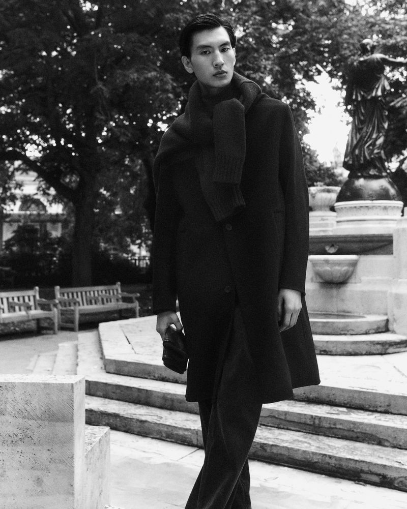 Monochrome portrait of a stylish Asian male model in an overcoat and scarf, posing on steps near benches and greenery in an outdoor setting.