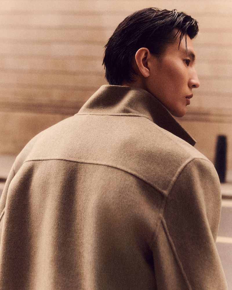 Profile view of an adult male wearing a tan collared coat, showcasing fashion style with neutral tones and soft lighting.