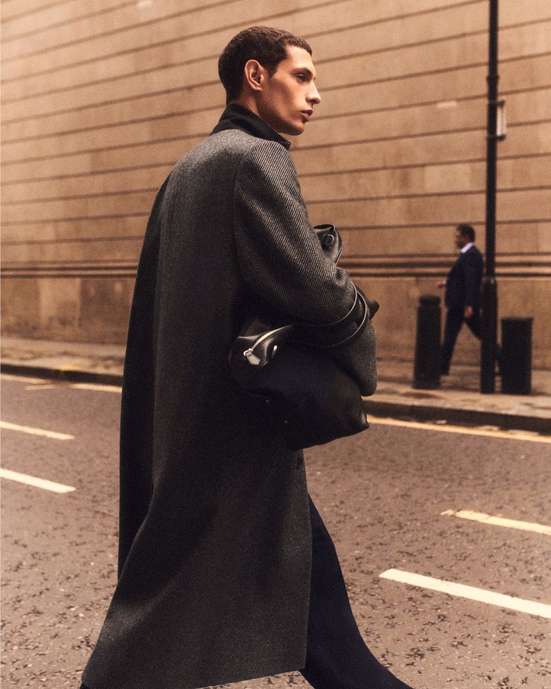 A man walks across a city street in a dark grey coat and holds a black bag, with a background view of buildings on a sunny day.