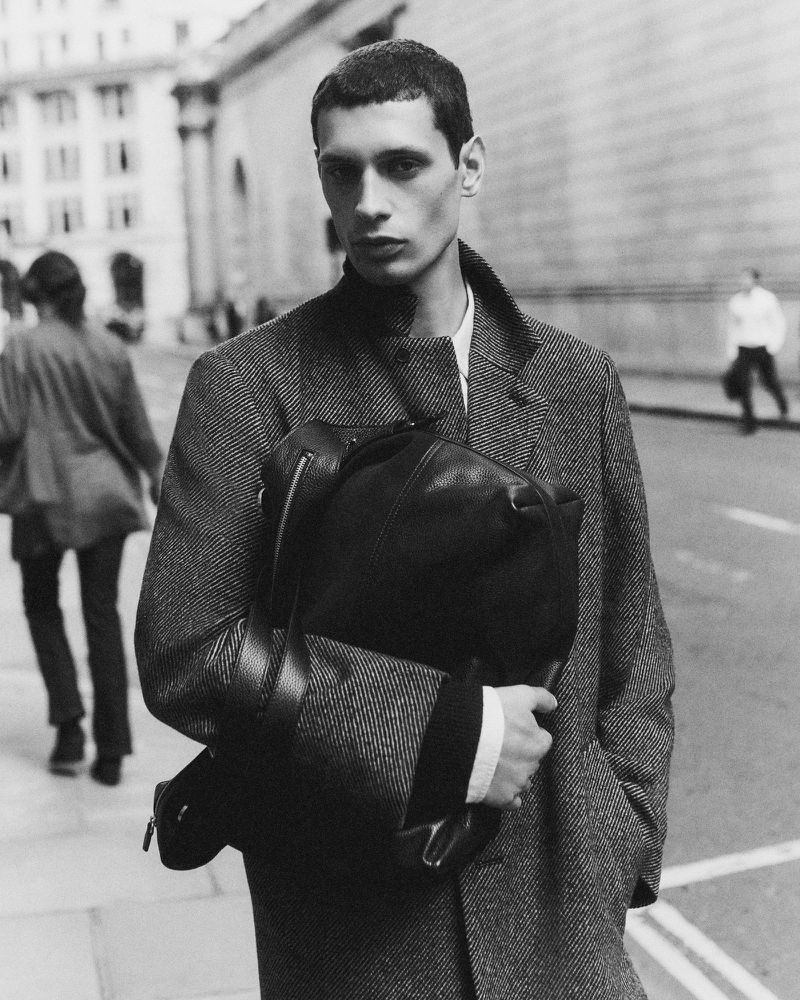 A monochrome portrait of a male model in a herringbone coat, holding a black leather backpack on an urban street with pedestrians.