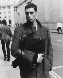 Black and white portrait of a man in a textured wool overcoat holding a black leather backpack while standing on a city street.
