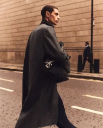 Model Tommaso Zana walking across an urban street in a long grey wool overcoat while carrying a black leather bag.