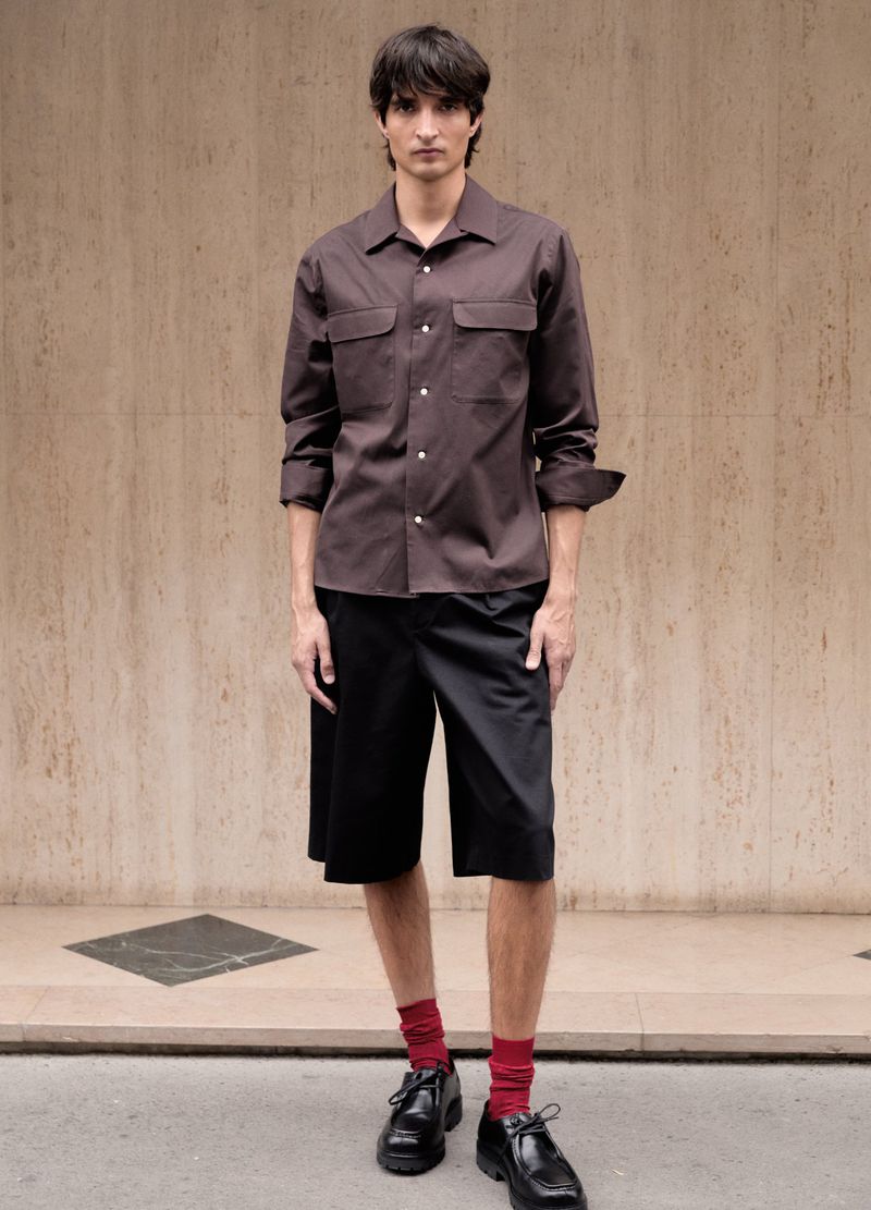 Frontal medium shot of a young adult male wearing a brown collared shirt and black shorts with red socks and black dress shoes.