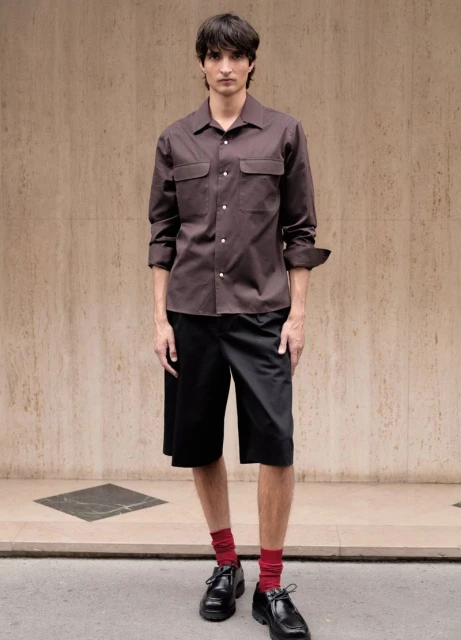 Frontal medium shot of a young adult male wearing a brown collared shirt and black shorts with red socks and black dress shoes.