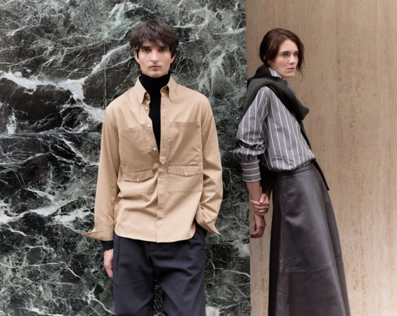 Man in beige utility shirt and black turtleneck standing beside a woman in a striped shirt and dark skirt against marble and stone pillars.