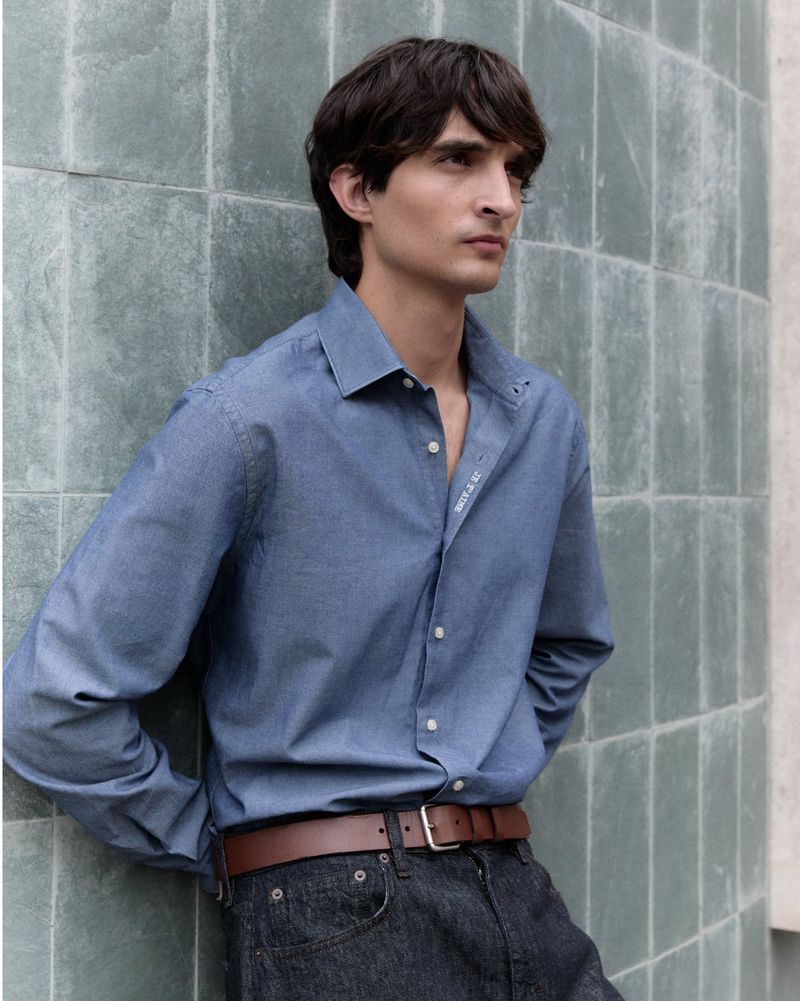 Male model leans against a tiled wall wearing a button-down shirt, brown leather belt, and jeans in a daytime fashion editorial.