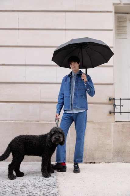 Man in denim outfit walks a black curly-haired dog under a black umbrella against a light stone wall backdrop in an urban setting.