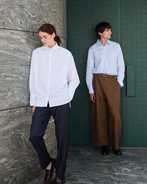 Two adult models, a male and a female, pose in dress shirts and trousers in an outdoor setting with a green door and stone wall.
