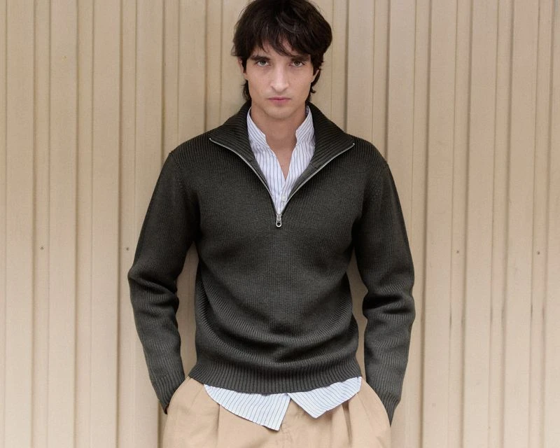 Man in olive sweater and khaki pants stands indoors against a light brown paneled wall. Portrait shot, medium framing, eye-level.