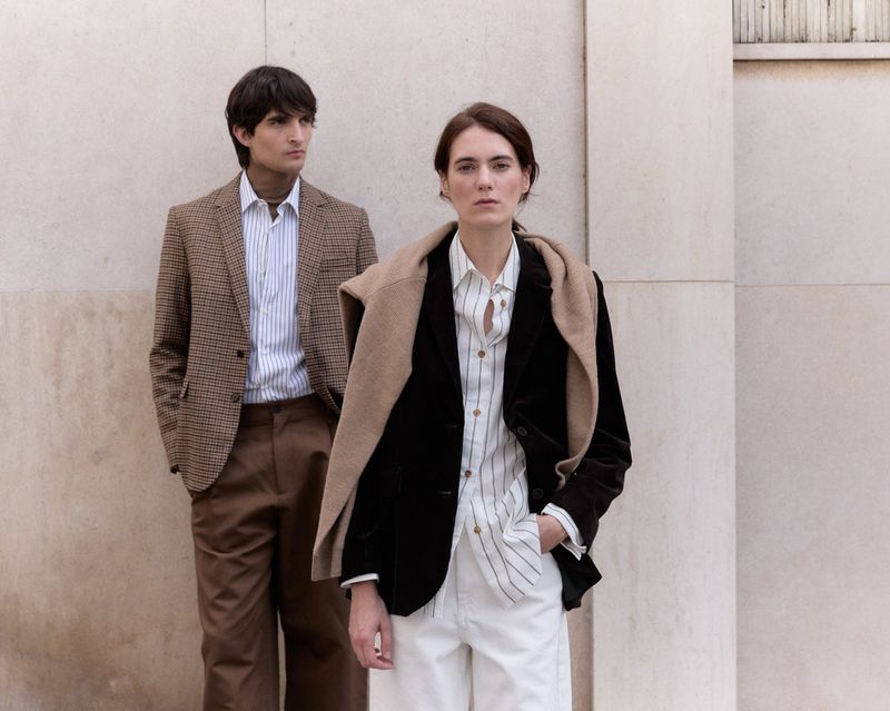 Two fashion models, one male and one female, pose in blazers and button-down shirts against a neutral wall for a fashion editorial.