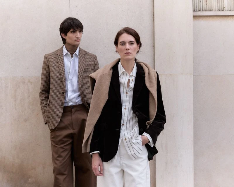 Two fashion models, one male and one female, pose in blazers and button-down shirts against a neutral wall for a fashion editorial.