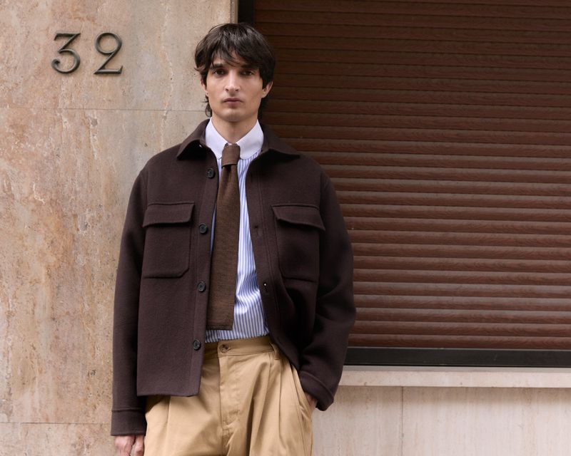 Adult male in a striped shirt, tie, and brown jacket stands outside against a brown wall. The scene is brightly lit.