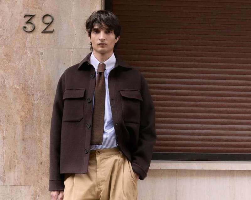 Adult male in a striped shirt, tie, and brown jacket stands outside against a brown wall. The scene is brightly lit.
