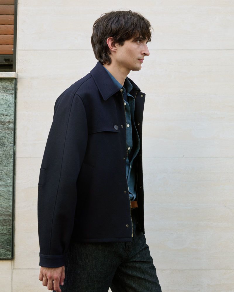 A man is posing outside wearing a navy jacket with a button-down shirt. The outdoor natural lighting enhances the colors of the garment.