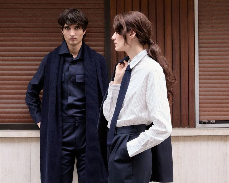 Two fashion models in stylish, predominantly navy blue business casual outfits pose outdoors in front of a wooden window shutter wall.