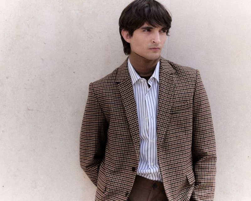 Portrait of a young adult man wearing a patterned blazer and turtleneck, posing against a textured wall in an outdoor setting.
