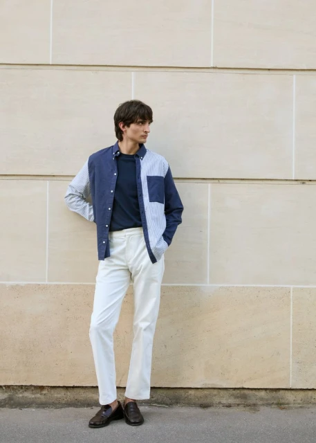 Man in casual fashion posing against a textured wall, wearing a colour-blocked shirt, white pants, and brown loafers.