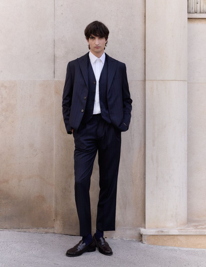A young adult male stands outdoors against a neutral wall, wearing a three-piece dark blue suit, white collared shirt, and dark brown loafers.