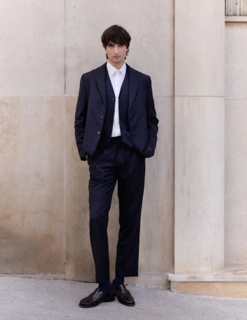 A young adult male stands outdoors against a neutral wall, wearing a three-piece dark blue suit, white collared shirt, and dark brown loafers.