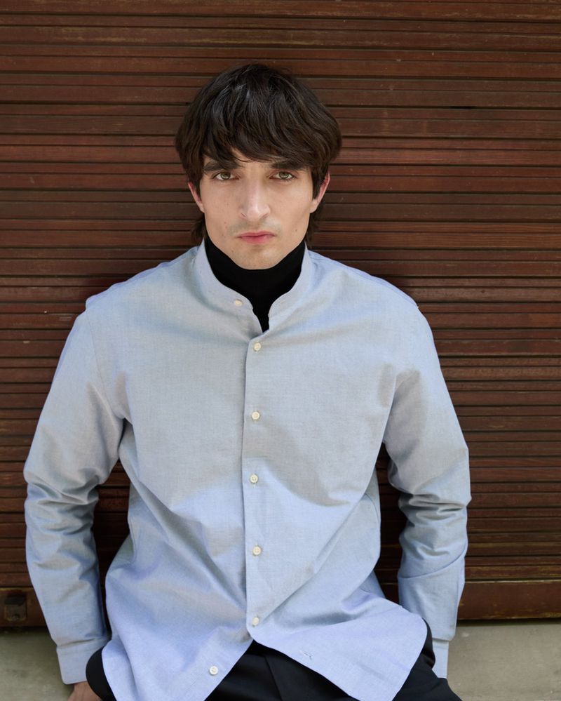 Portrait of a dark-haired adult male in a blue button-down shirt and black turtleneck, posing in front of a wooden shutter backdrop.
