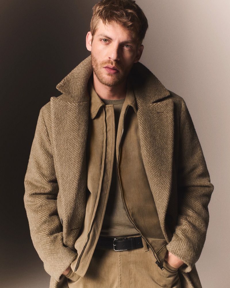 A blond adult male poses in a tan herringbone overcoat, jacket, and corduroy pants. The lighting is studio with a two-tone background.