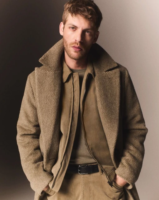 A blond adult male poses in a tan herringbone overcoat, jacket, and corduroy pants. The lighting is studio with a two-tone background.