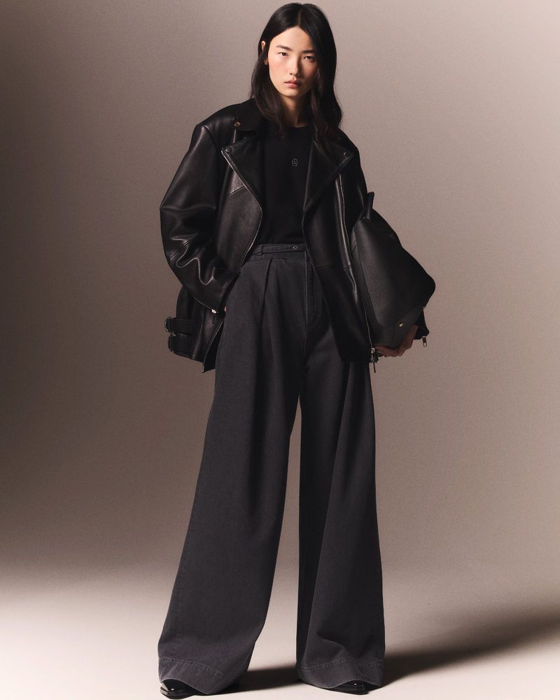 A young Asian woman models black leather jacket, black shirt, and wide-leg pants in a studio with diffused lighting.