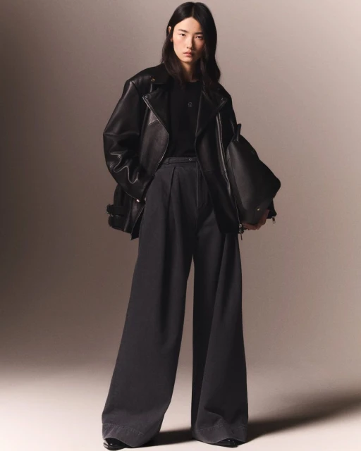 A young Asian woman models black leather jacket, black shirt, and wide-leg pants in a studio with diffused lighting.