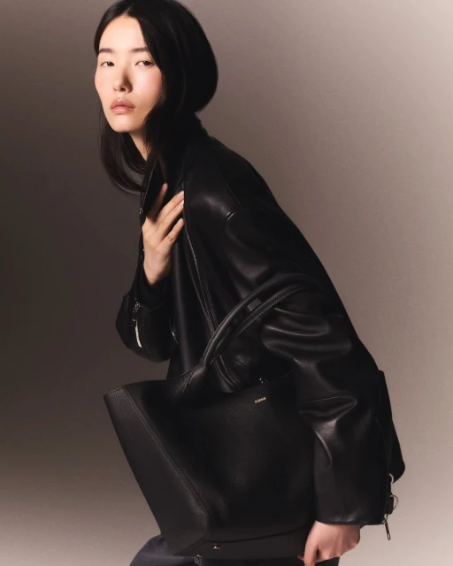 Elegant Asian woman in leather jacket poses with a black tote bag against a neutral backdrop in a fashion forward studio shot.