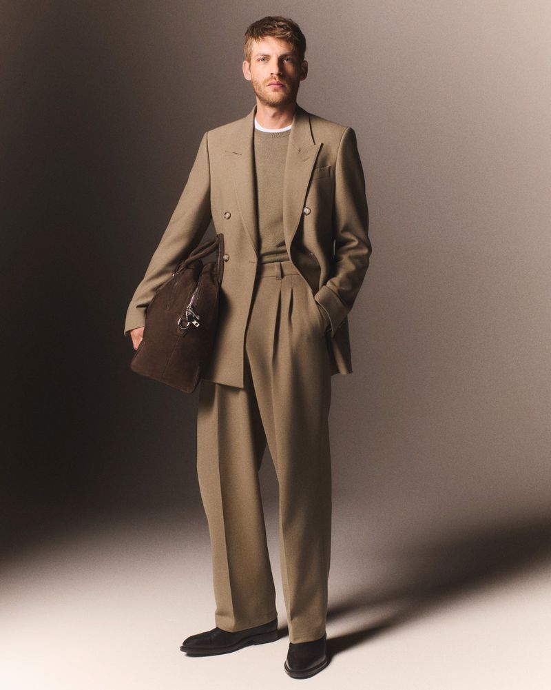 Male fashion model in a tan suit and brown shoes, holding a brown leather bag, posing in a studio with a neutral grey background.