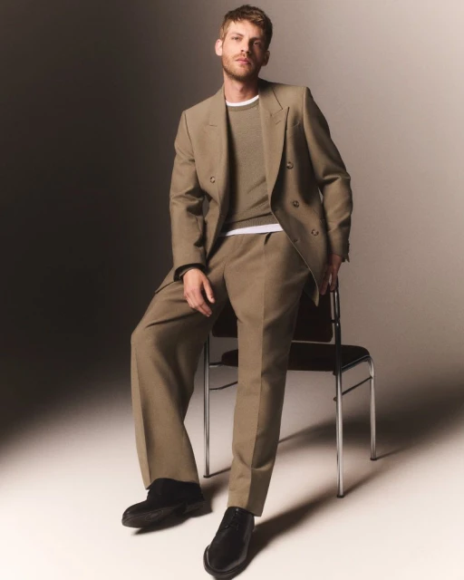 Man in a tan suit seated on a chair. He wears a crewneck sweater and black shoes. The shot is lit by studio lighting.