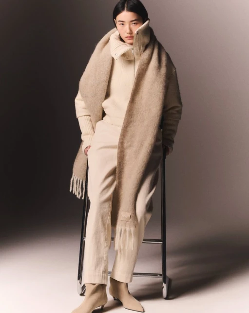 Fashion portrait of an Asian adult female wearing a sweater, pants, boots, and a long scarf while sitting on a metal stool.