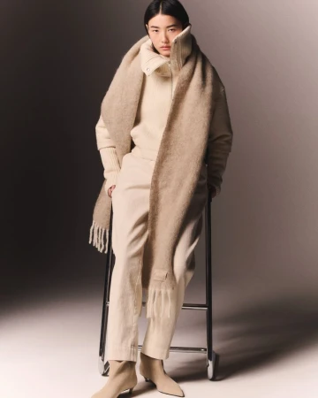 Fashion portrait of an Asian adult female wearing a sweater, pants, boots, and a long scarf while sitting on a metal stool.
