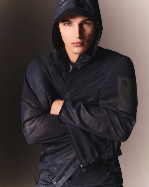 Close-up of a fair-skinned adult male wearing a hooded black windbreaker with arms crossed against a neutral grey studio backdrop.