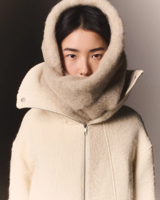 A woman with light skin and dark hair is the primary focus, wearing a white wool coat and a large beige knitted scarf covering her head.