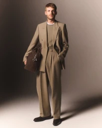 Man wearing a tailored taupe double-breasted suit holding a dark brown leather weekender bag against a neutral studio backdrop