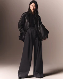 Model in oversized black leather jacket and wide grey trousers holding a black handbag against a gradient backdrop