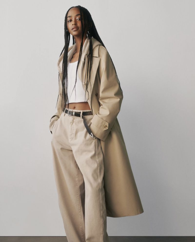 A slender young Black woman with braided hair poses in a studio, wearing a white top, baggy beige pants, black belt, and tan trench coat.