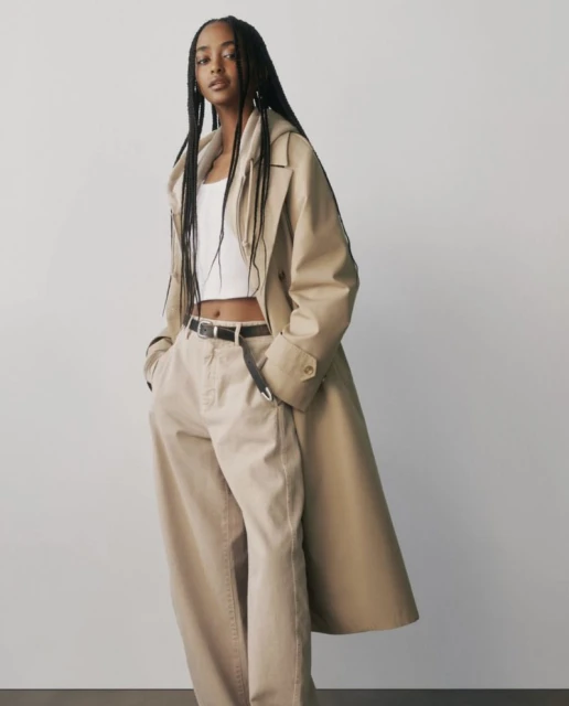 A slender young Black woman with braided hair poses in a studio, wearing a white top, baggy beige pants, black belt, and tan trench coat.