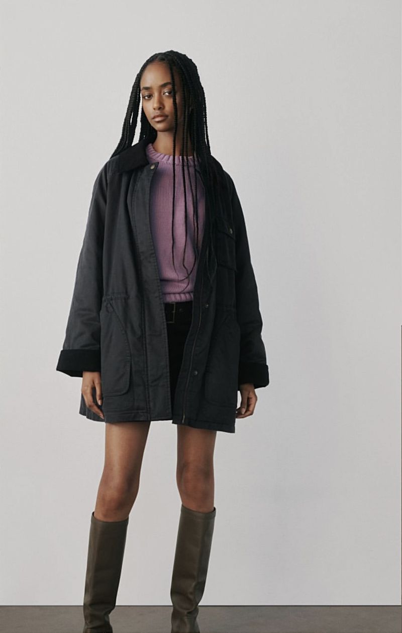 A young woman in a lavender sweater, black jacket and olive boots poses against a white backdrop in a full studio shot.