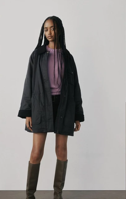 A young woman in a lavender sweater, black jacket and olive boots poses against a white backdrop in a full studio shot.