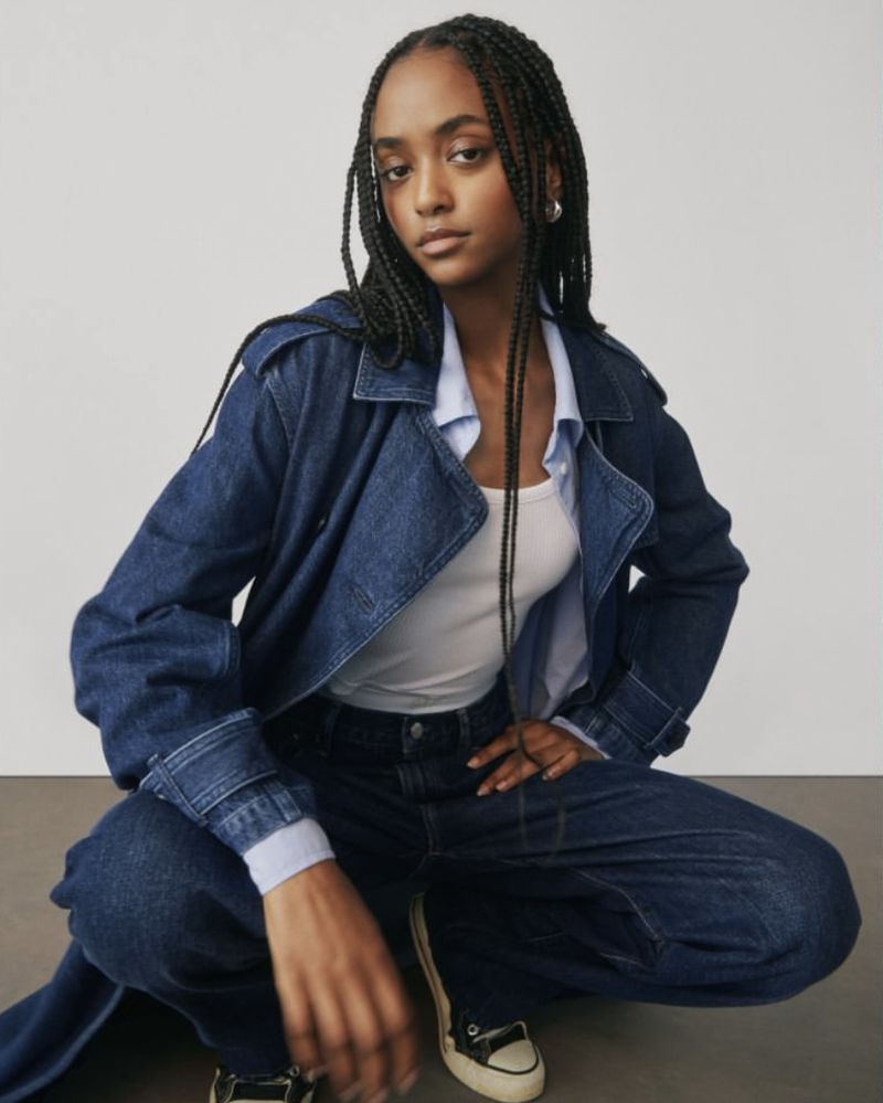 A young Black woman crouches indoors wearing a denim jacket, light blue shirt, white tank top, dark jeans, silver earrings, and sneakers.