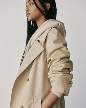 Model with braided hair wearing a beige trench coat layered over a hoodie against a plain white studio background.